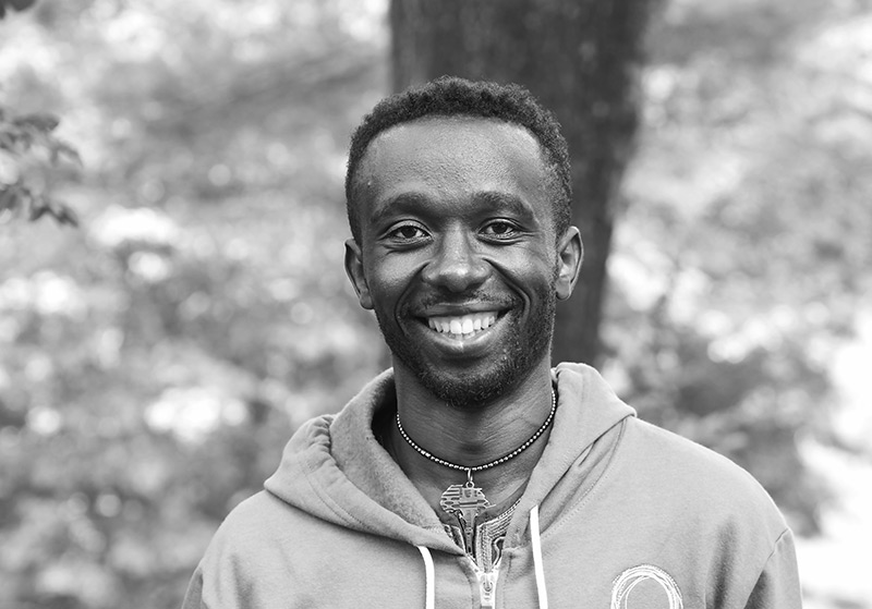 Black and white portrait of Sunday Nzititira. He has short black hair and is wearing a World Learning Sweater with the green circle logo, grinning widely