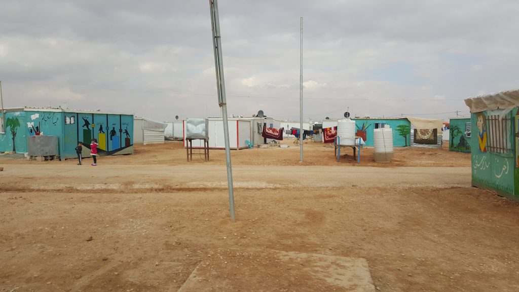 Small blue and white buildings, some with canvas tarps attached, on a dry, flat landscape.