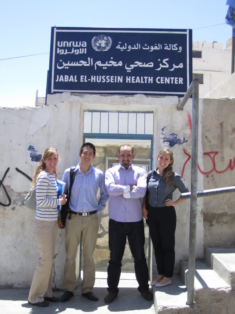 Three students stand with a male doctor in front of a doorway. The sign above says "Jabal El-Hussein Health Center."