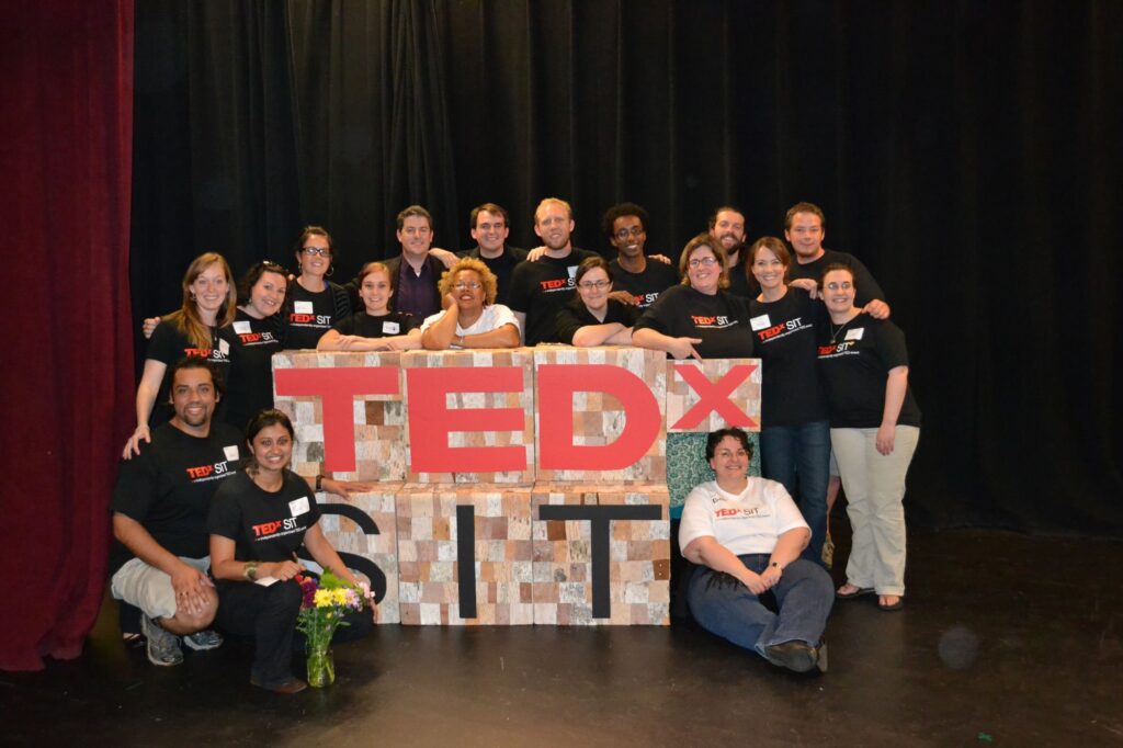 Eighteen people on a stage pose around a sign that says "TEDx SIT."