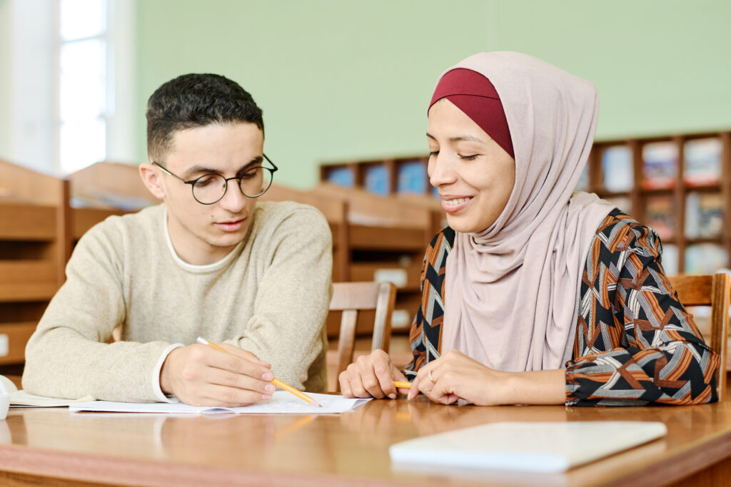 Young man and woman in hijab sitting at desk in classroom reading papers.