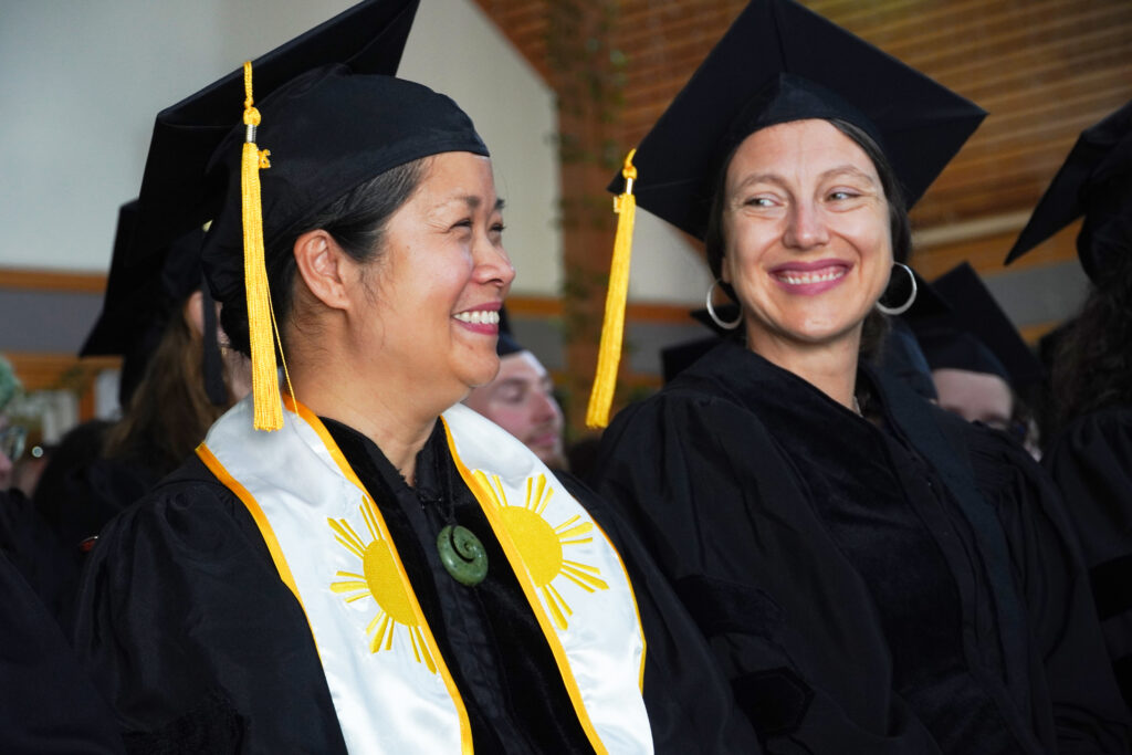 Two women are smiling at each other. Both are wearing black graduation robes and caps.