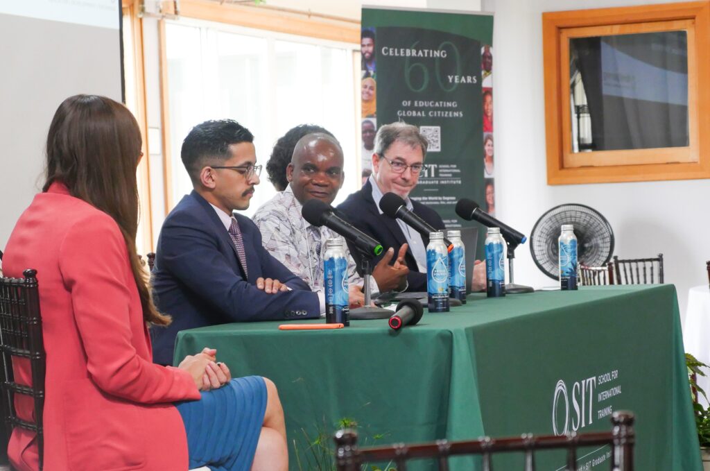 A panel of four people sit at a table with the SIT logo. Microphones are on the table, and a woman sits to the left of the table listening to them speak.