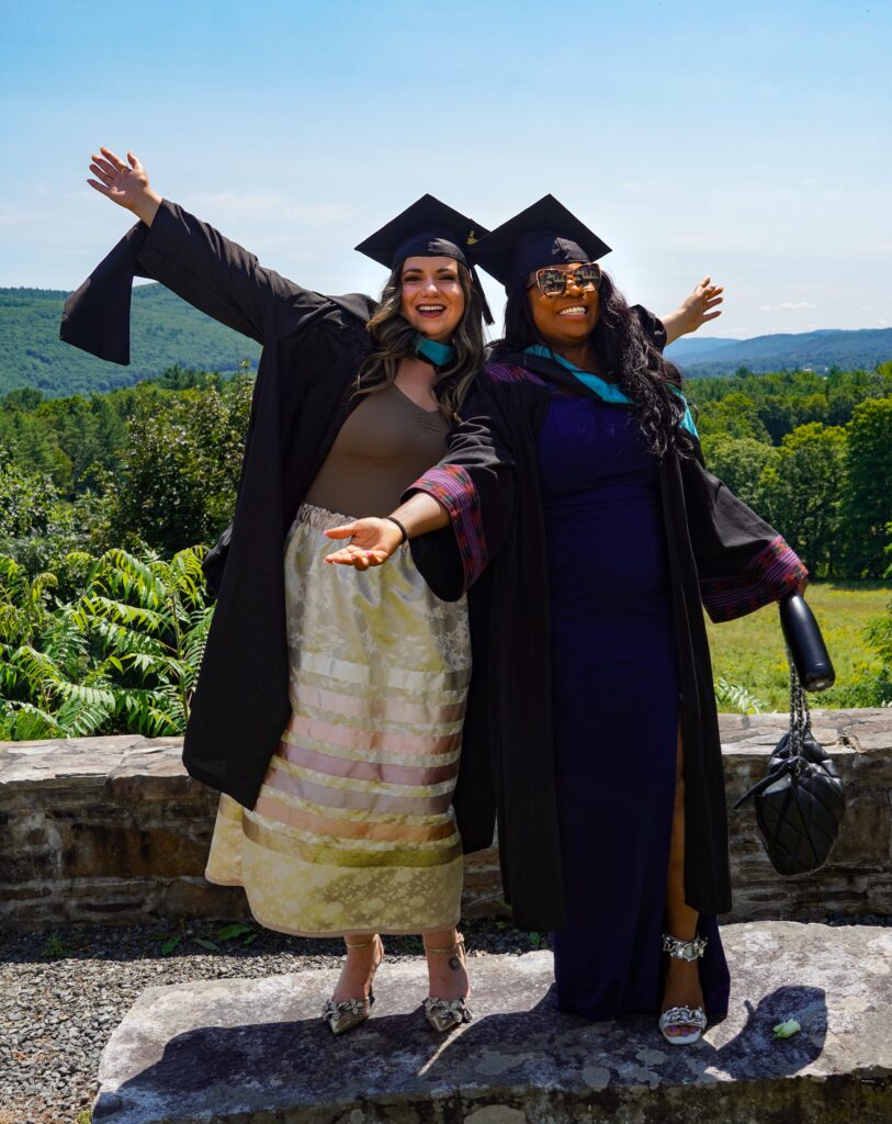 Two women pose with arms stretched out, wearing graduation caps and gowns. They are standing on a rock and green hills are behind them.