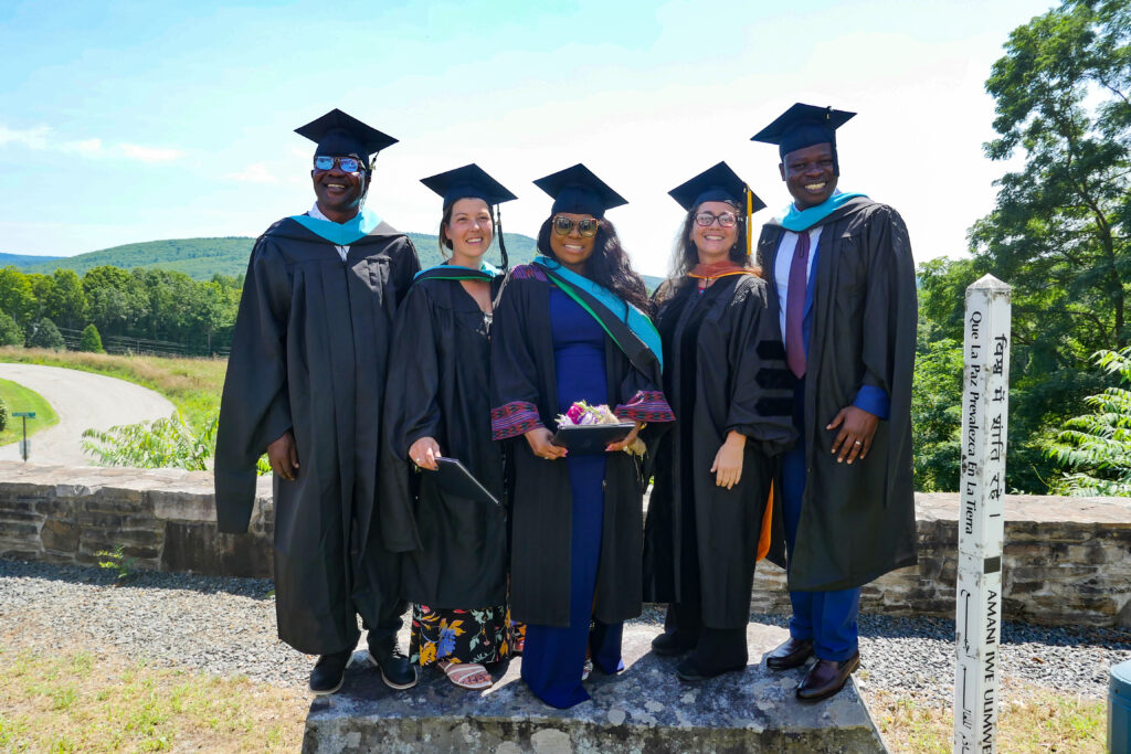 Three women and two men pose on a rock. All wear black graduation robes and caps and behind them is a stone wall and trees and road leading away.