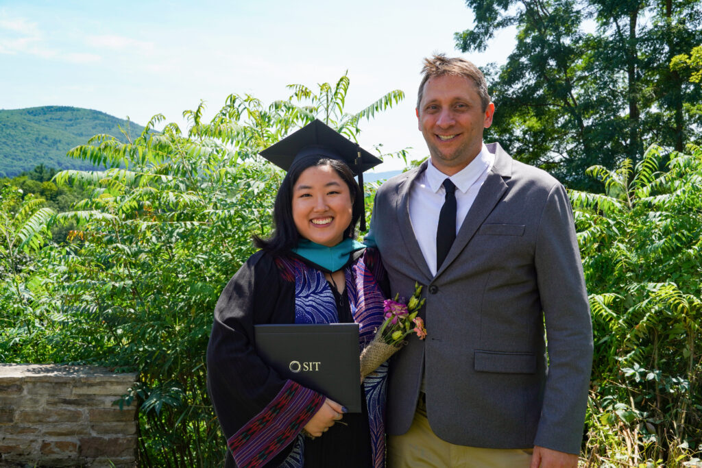 A women in a black graduation robe and cap poses next too a man wearing a suit coat and tie. They are both smiling and trees are behind them.