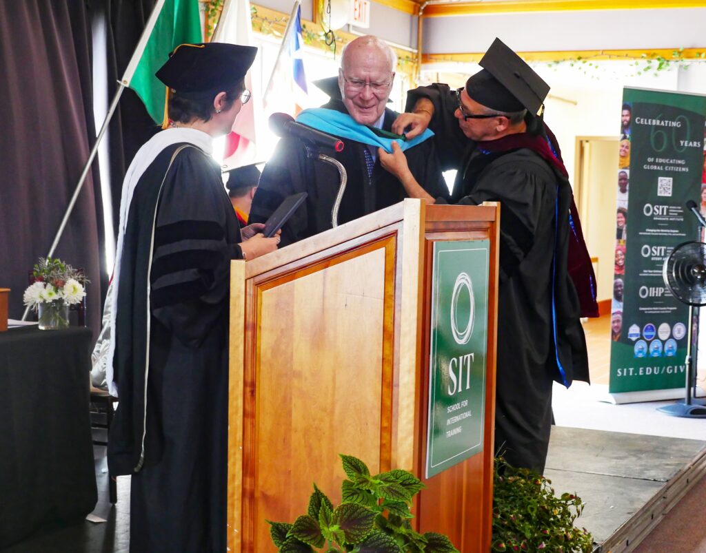 A woman and a man are pinning a graduation hood onto a man standing in the middle of them. All are wearing graduation robes and a podium is in front of them.