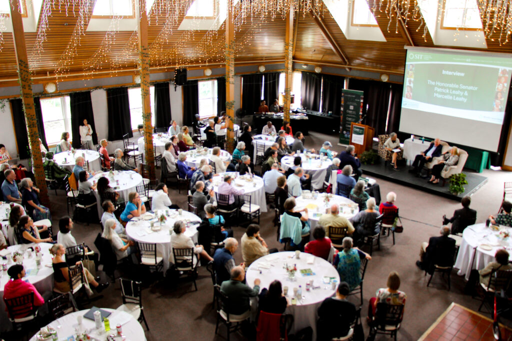 Overhead view of a large room full of round tables. Conference guests are seated at the tables and facing the front of the room with a stage and large projected screen.