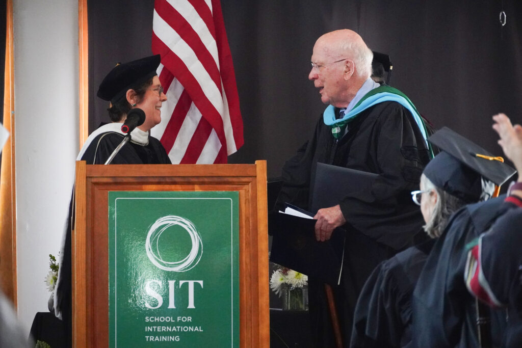 A woman and a man greet each other, shaking hands, behind a podium. Both are wearing black graduation robes. An American flag is in the background.