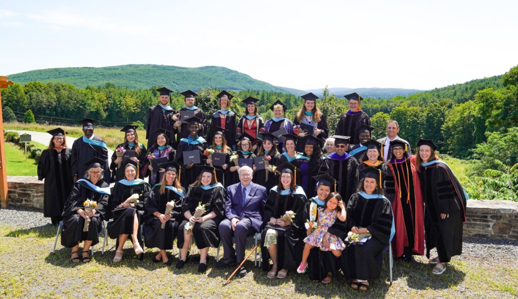 Large group of adults pose in three rows wearing graduation caps and gowns. A stone wall is behind them the the view shows green hills in the background.