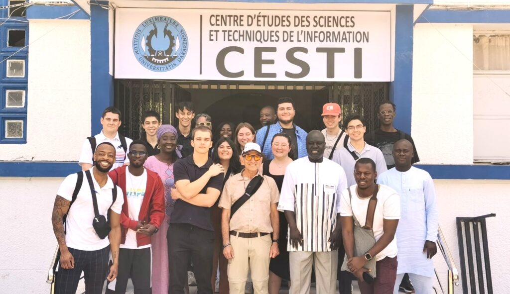 Two rows of college aged students are standing under a large sign for a university in Senegal. They are posing with several adults.