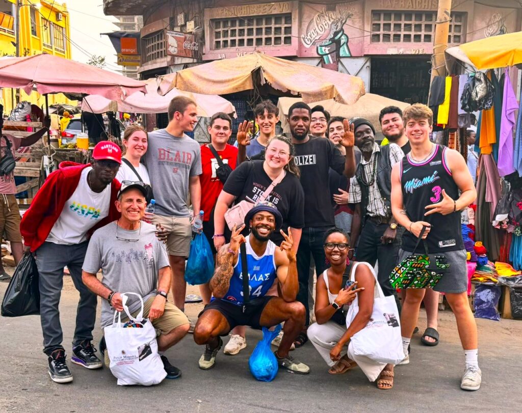 A group of approximately 15 college age students and some adults pose in front of market stalls with large umbrellas. 