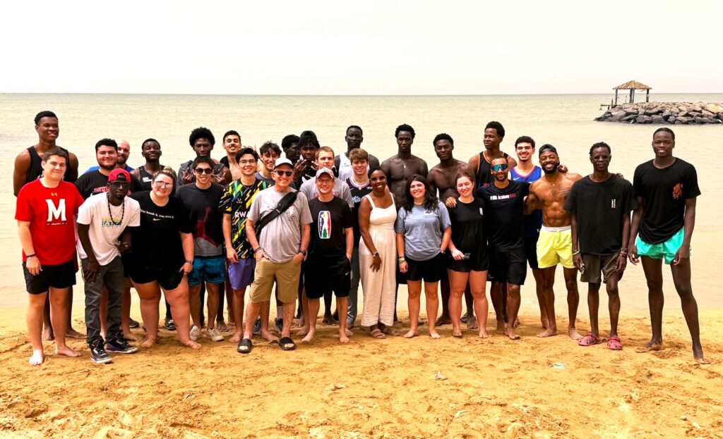 More than 25 college aged young adults are standing on a beach. There is an ocean behind them and and pier made of stone.