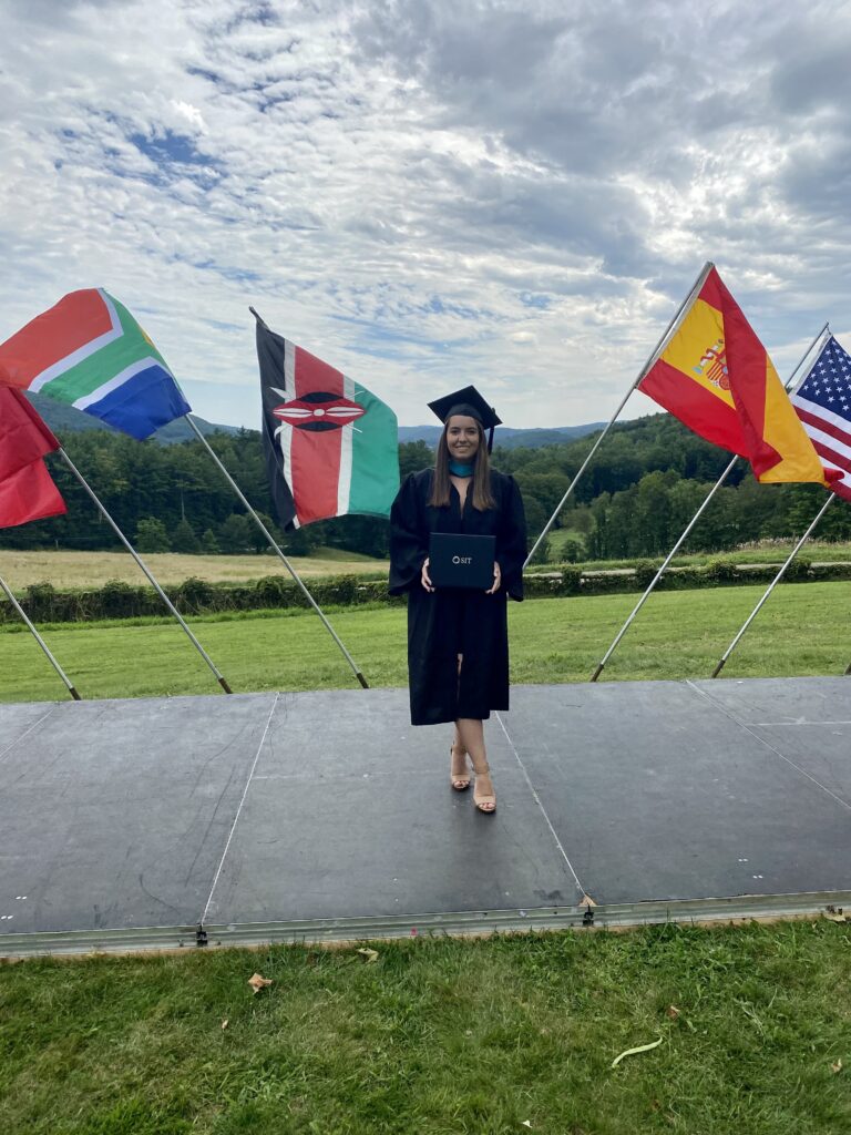 A young adult wearing a graduation cap and gown holds a degree in front of an outdoor graduation ceremony space with international flags and a scenic mountain background.