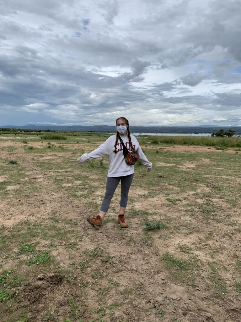 A young adult wearing an Elon University sweater and a face mask poses in a Ugandan field.