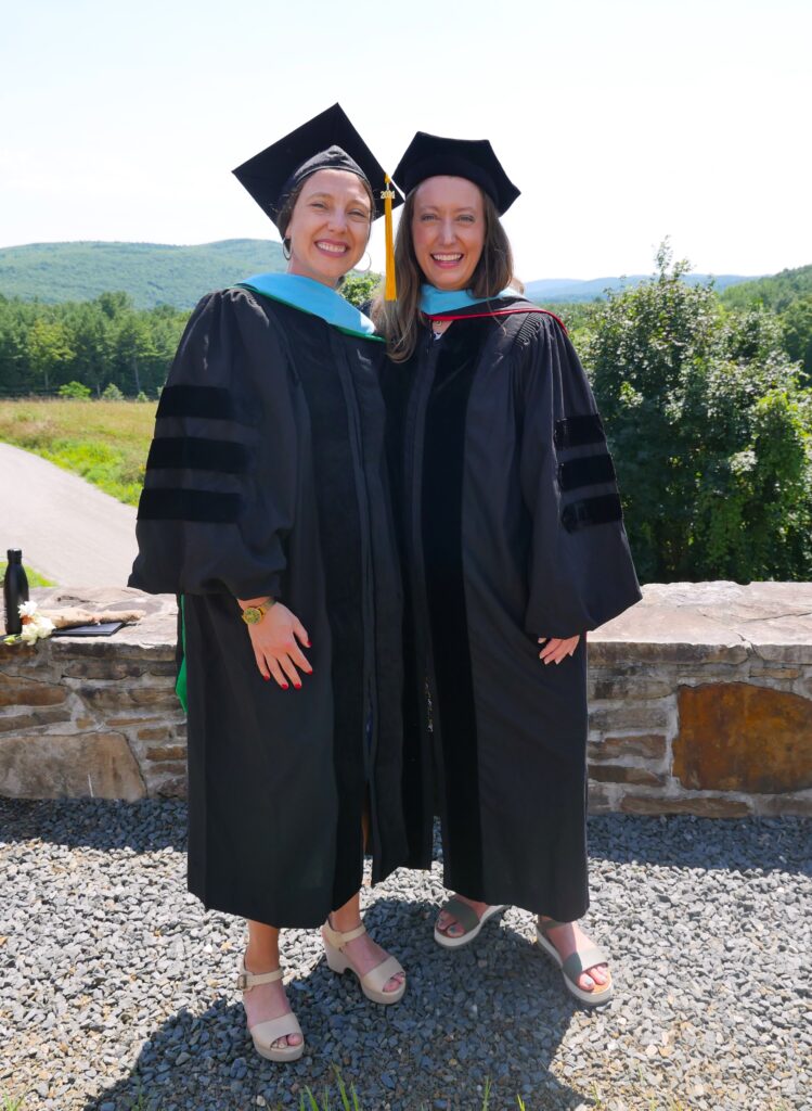 Two women standing, both wearing black graduation gowns and caps. Behind them is a stone wall and then green hills.