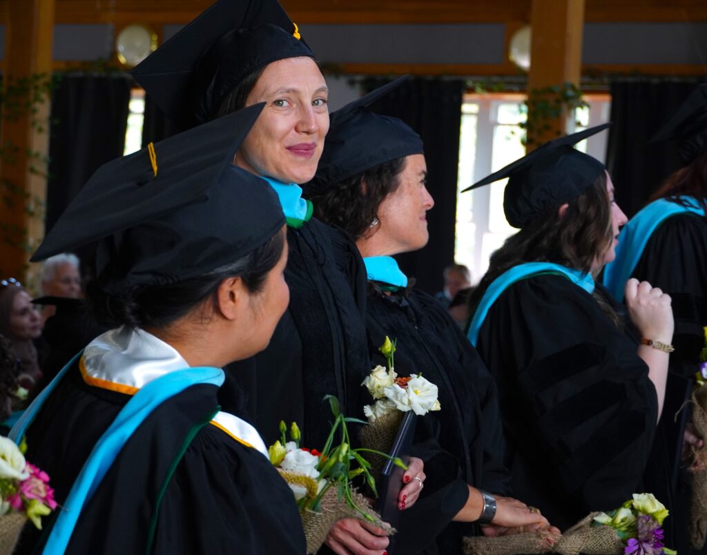 Four women stand in a row, wearing black graduation gowns and caps. One woman has her head turned and is looking at the camera. All others are looking ahead.