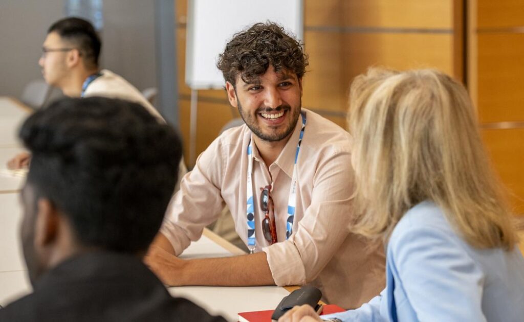 Young man with curly brown hair sitting at a conference table. He is turned and talking to the woman next to him.